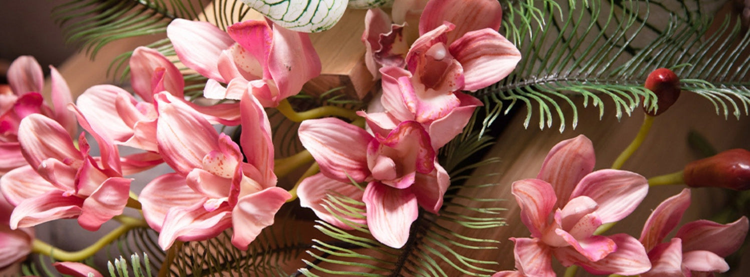 Rózsaszín mű orchidea virágok Real Touch hatással, részletgazdag kidolgozással, dekoratív zöld levelekkel, természetes hatású kompozícióban.