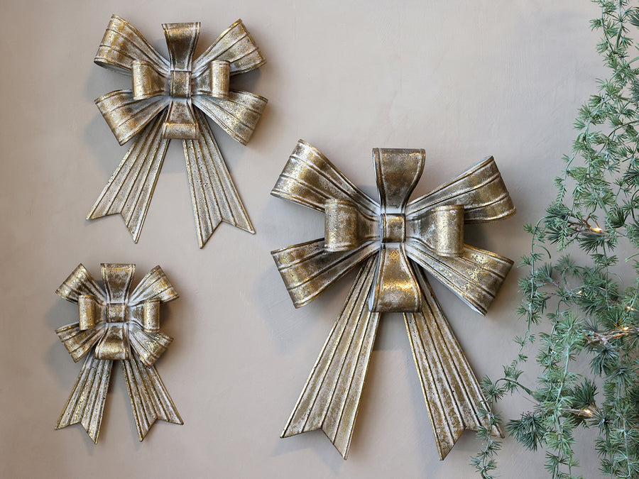Three antique gold metal ribbon bows in varying sizes mounted on a neutral wall.