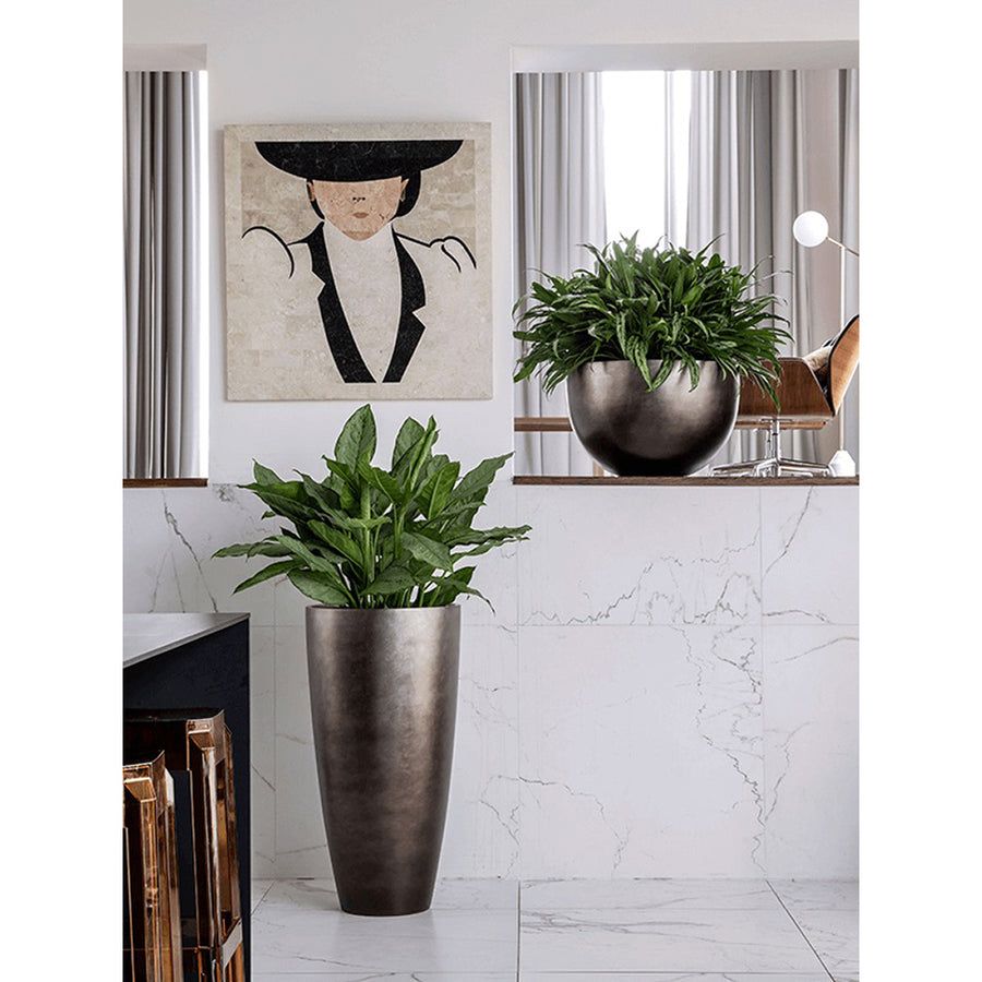 Modern enteriőrben elhelyezett BAQ METALLIC BOWL matt barna kaspó zöld növényekkel, márványhatású fal előtt, dekorációként.