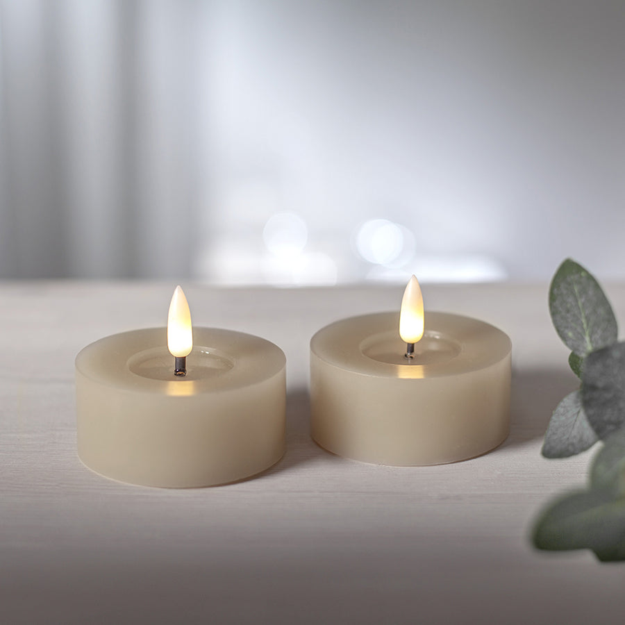 Két beige színű LED teamécses meleg fehér fénnyel világítva, világos fa felületen, háttérben bokeh fényekkel.
