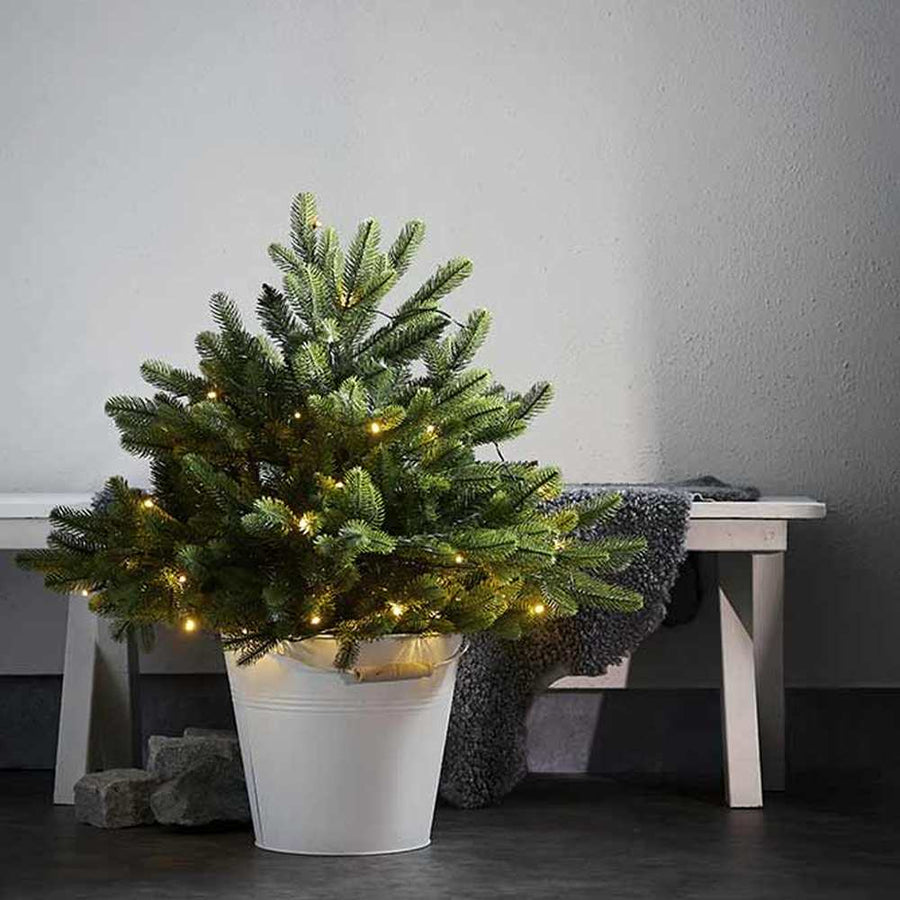 Mesterséges, 80 cm átmérőjű és 75 cm magas, kül és beltérben is egyaránt használható, 40 db meleg fehér fényű, időzítővel ellátott LED világítással felszerelt, élethű dekorációs betűzős műfenyő fehér zománcozott vödörben, fehér pad előtt, szürke padlón