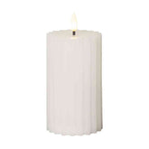 Fehér, valódi viaszból készült LED gyertya 15 cm magas, kannelúrás oszlopdizájnnal és meleg fényű pislákoló lánggal, elegáns dekorációhoz.
