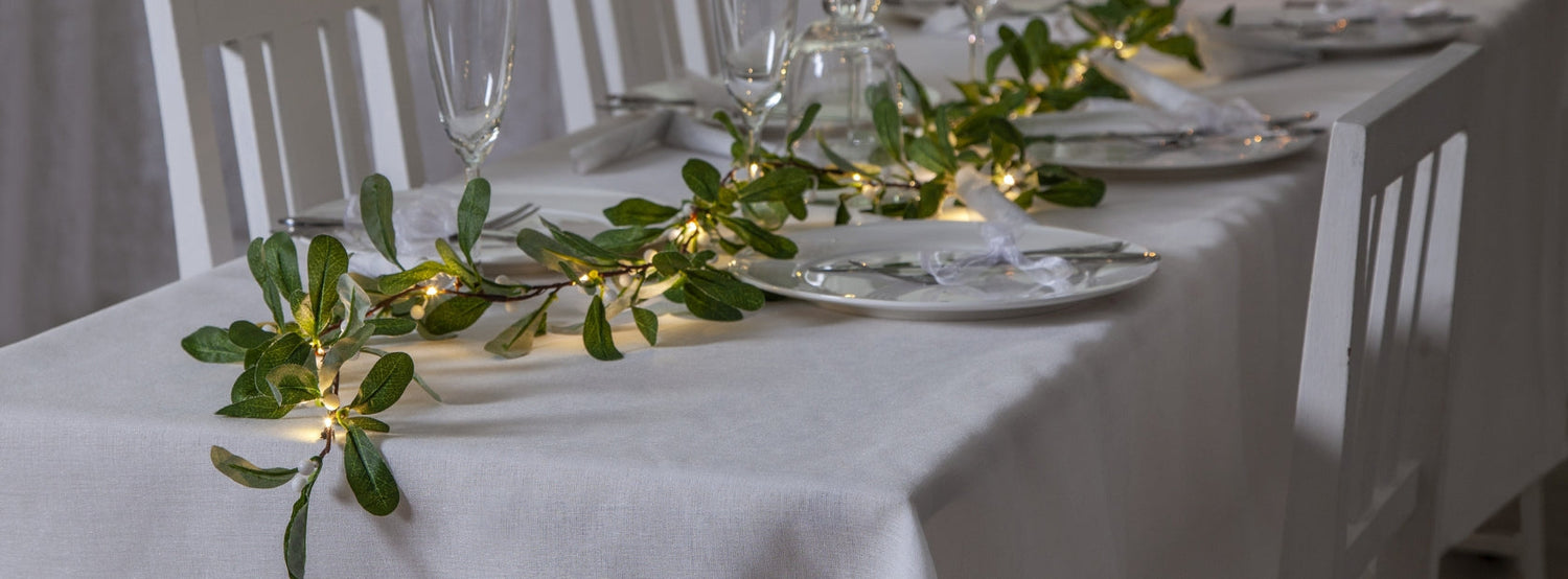 Világító műnövény girland melegfehér LED fényekkel megvilágítva, elegánsan végigfut egy ünnepi asztalon, fehér abrosz és stílusosan terített tányérok között, elegáns poharakkal és fehér szalvétákkal – hangulatos beltéri dekorációs megoldás.