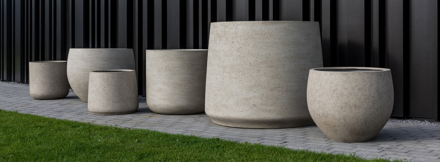 Pottery Pots Urban kollekció kaspói kültéren, ficonstone anyagból készült betonhatású óriás kaspók fekete fal előtt elrendezve.