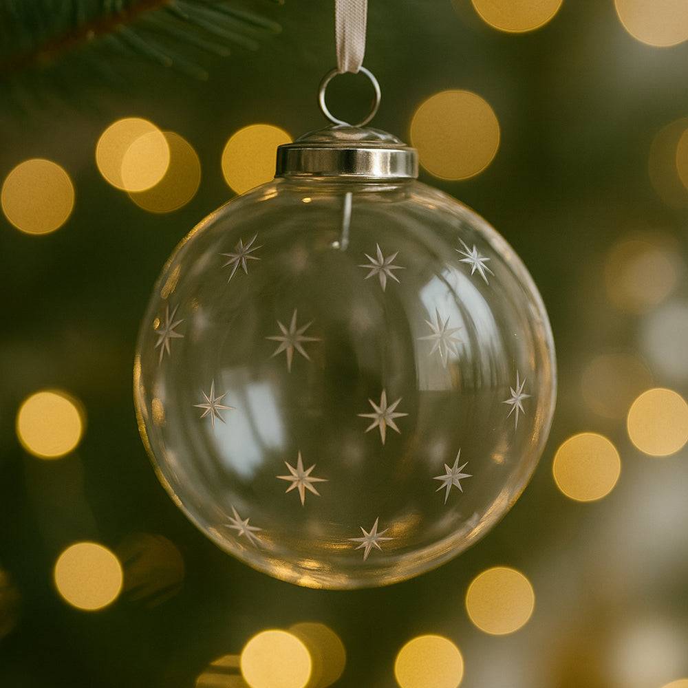 Karácsonyfán függő áttetsző üveggömb, maratott csillagmintákkal és ünnepi fényekkel a háttérben.