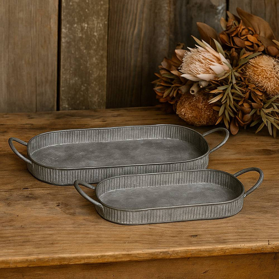 Két kézzel készült, antikolt felületű szürke fém tálca füllel, rusztikus fa felületen, száraz virágkompozícióval a háttérben, jobbról érkező meleg fényben megvilágítva.