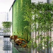 Modern kültéri térben elhelyezett 200 cm magas, UV-álló mű bambusz térelválasztó fekete fa kaspóban, zöld növényfallal a háttérben.