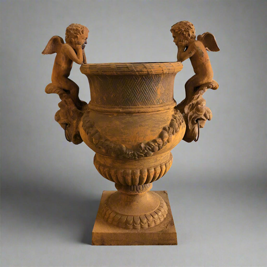 Château Jardin antikolt öntöttvas kaspó puttófigurákkal, műtermi világításban, klasszikus díszítőelemekkel és patinás felülettel.