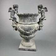 Château Jardin antikolt öntöttvas kaspó puttófigurákkal, műtermi világításban, klasszikus díszítőelemekkel és patinás felülettel.