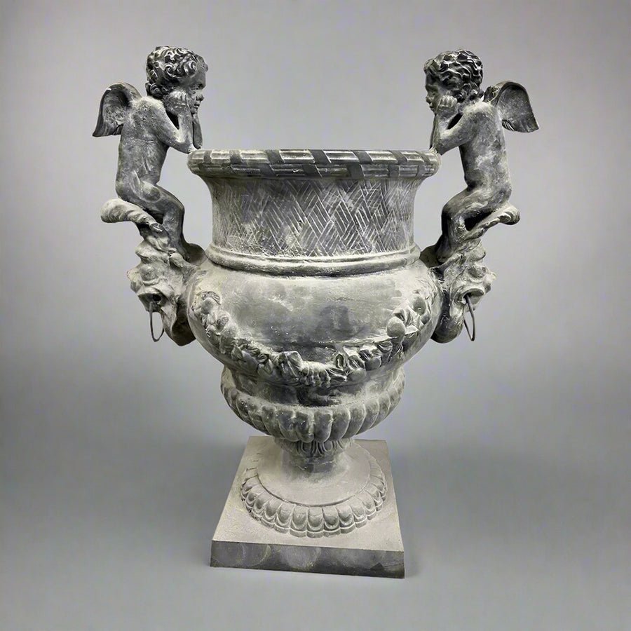 Château Jardin antikolt öntöttvas kaspó puttófigurákkal, műtermi világításban, klasszikus díszítőelemekkel és patinás felülettel.