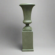 Antik zöld, nyújtott formavilágú kaspó letisztult posztamensen, stúdió környezetben, semleges szürke háttérrel.