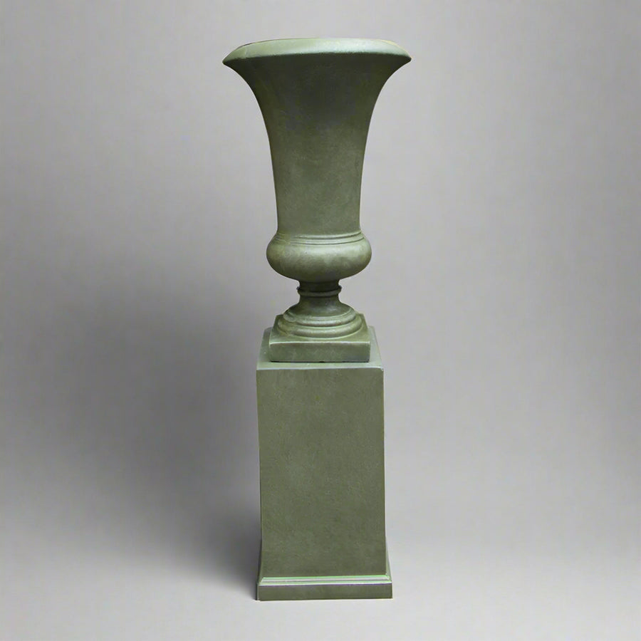 Antik zöld, nyújtott formavilágú kaspó letisztult posztamensen, stúdió környezetben, semleges szürke háttérrel.