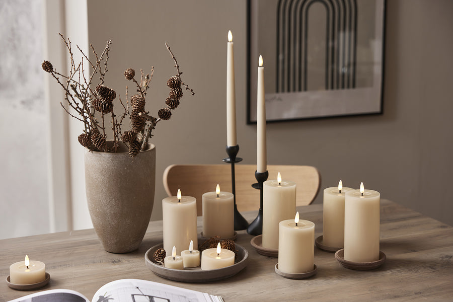 Bézs LED gyertyák különböző méretben és stílusban tálcán és gyertyatartóban, természetes dekorációval berendezett étkezőasztalon.
