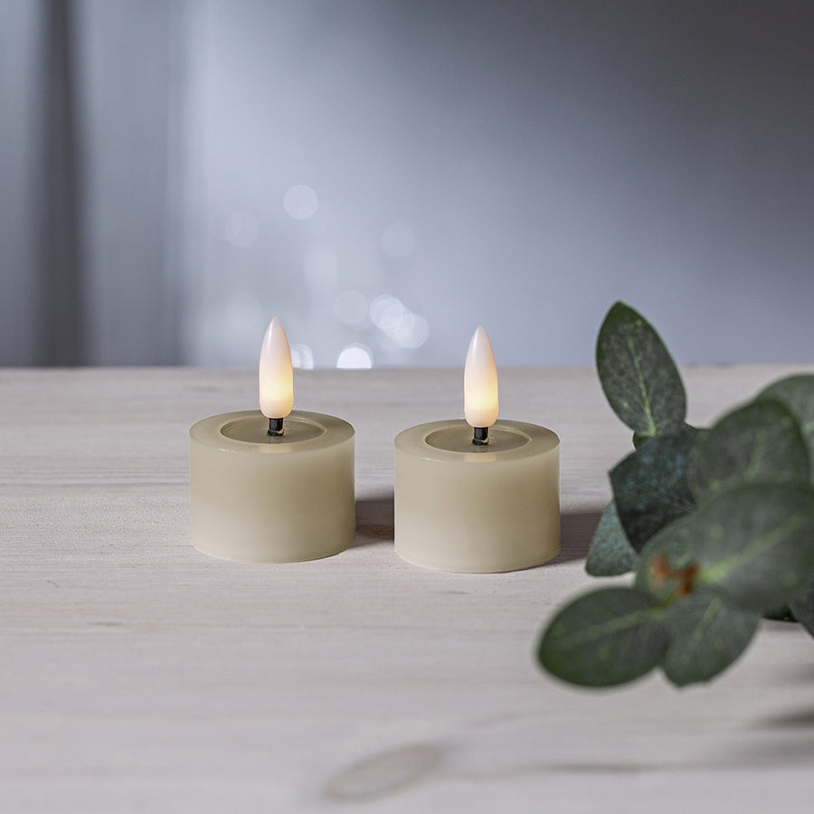 Két bézs színű LED teamécses világít világos faasztalon, elmosódott háttérrel és zöld levél dekorációval.