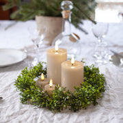 Bézs LED teamécsesek és henger gyertyák zöld koszorúval díszített ünnepi asztalon, háttérben elegáns teríték és üvegkancsó.