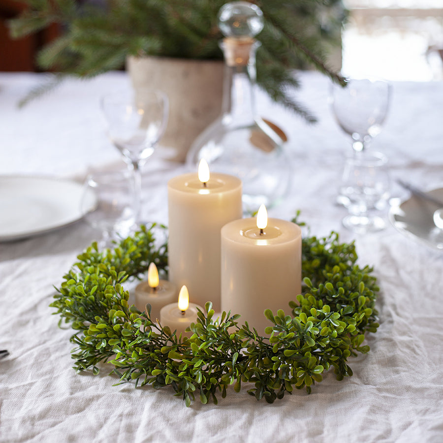 Bézs LED teamécsesek és henger gyertyák zöld koszorúval díszített ünnepi asztalon, háttérben elegáns teríték és üvegkancsó.