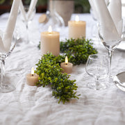 Bézs LED teamécsesek és gyertyák ünnepi asztalon, fehér terítővel, zöld girlandokkal és elegáns poharakkal díszítve.