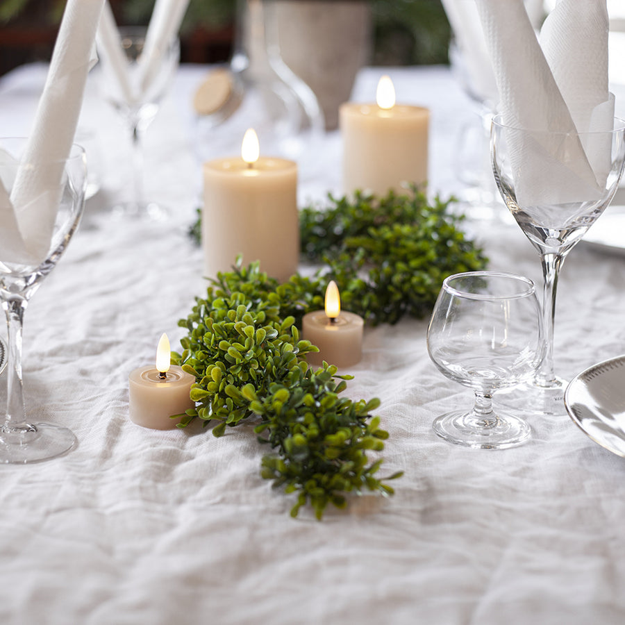 Bézs LED teamécsesek és gyertyák ünnepi asztalon, fehér terítővel, zöld girlandokkal és elegáns poharakkal díszítve.