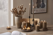 Szürkésbarna LED gyertyák különböző méretben, stílusos kompozícióban elrendezve fa asztalon, természetes dekorációval és elegáns háttérrel.
