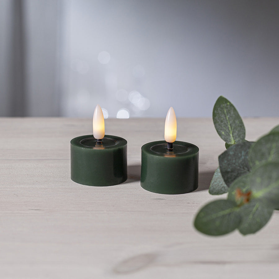 Két zöld LED teamécses meleg fényű lánggal, világos fa felületen elhelyezve, bokeh háttérrel és eukaliptusz ág dekorációval.