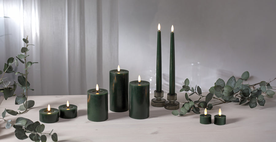 Két darab zöld Flow LED vacsoragyertya, oszlopgyertyák és teamécsesek társaságában, elegáns dekorációként eukaliptuszágakkal egy világos enteriőrben.