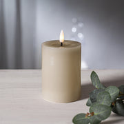 Bézs színű LED oszlopgyertya valódi viaszból, pislákoló lánggal, világos háttér előtt, dekoratív zöld eukaliptuszággal.