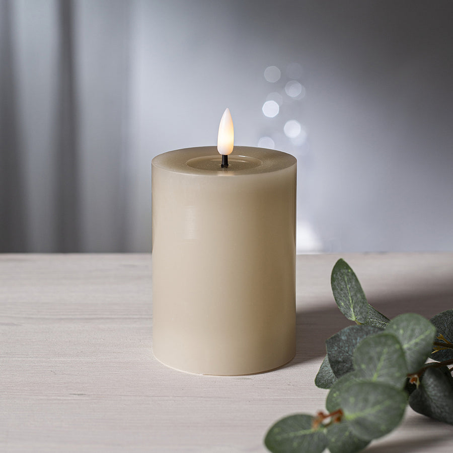 Bézs színű LED oszlopgyertya valódi viaszból, pislákoló lánggal, világos háttér előtt, dekoratív zöld eukaliptuszággal.