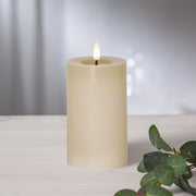 Bézs színű LED oszlopgyertya valódi viaszból, pislákoló lánggal, világos háttér előtt, dekoratív zöld eukaliptuszággal.
