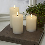 Fehér LED gyertyák a Star Trading Flow kollekcióból, természetes hatású lánggal, dekorációként elhelyezve egy könyvre, növénnyel és ablakpárkánnyal a háttérben,