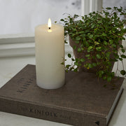 Fehér LED gyertya a Star Trading Flow kollekcióból, természetes hatású lánggal, dekorációként elhelyezve egy könyvre, növénnyel és ablakpárkánnyal a háttérben.