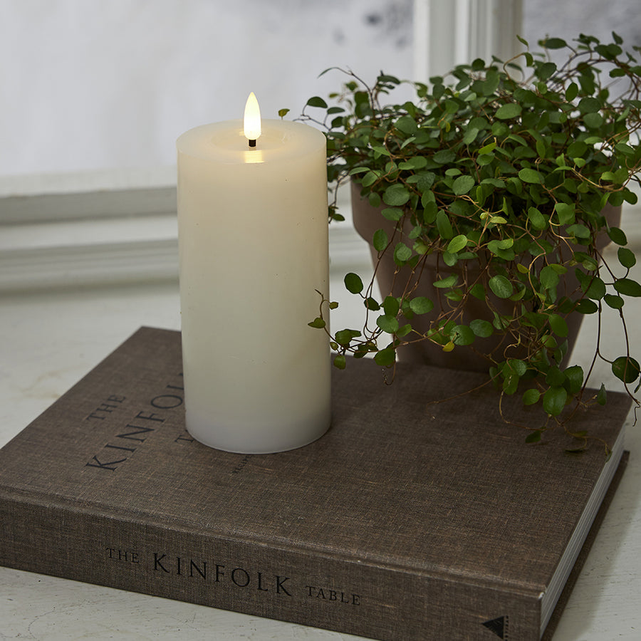 Fehér LED gyertya a Star Trading Flow kollekcióból, természetes hatású lánggal, dekorációként elhelyezve egy könyvre, növénnyel és ablakpárkánnyal a háttérben.