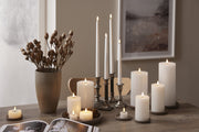 Fehér LED gyertyák különböző méretekben, elegáns tálcákon és gyertyatartókban elhelyezve egy természetes, letisztult stílusú asztaldísz részeként.