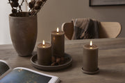 Három szürkésbarna LED gyertya világít egy fa asztalon, természetes tobozokkal és vázával kiegészítve, meghitt hangulatot árasztva.