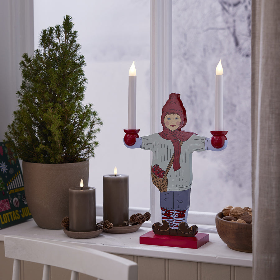 Két szürkésbarna LED gyertya egy ablakpárkányon, ünnepi dekorációval: fenyő, dió, és egy bájos figurás gyertyatartó hóval borított háttérrel.