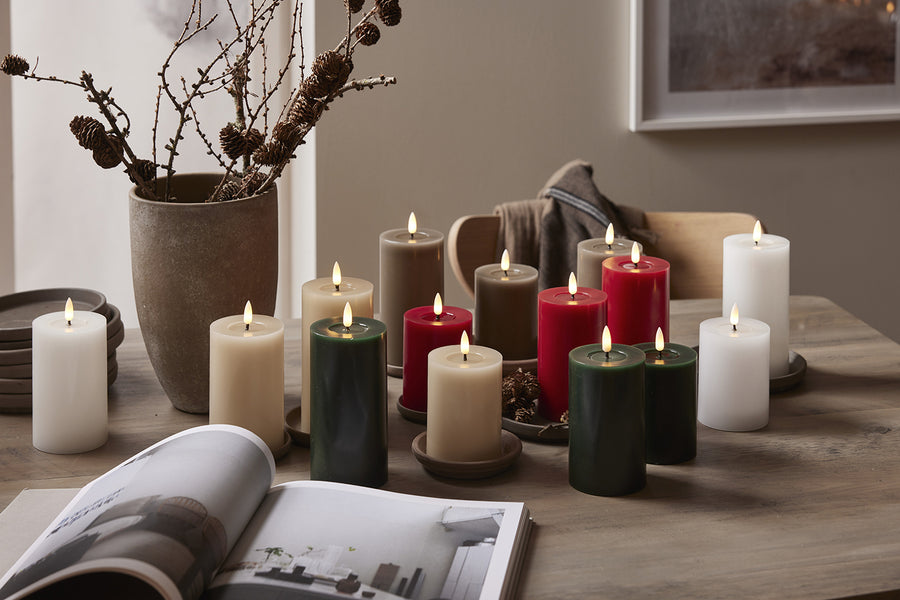 LED gyertyák különböző színekben – fehér, bézs, zöld, piros és szürkésbarna – ünnepi elrendezésben egy fa asztalon, természetes dekorációval.