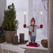 Két szürkésbarna LED gyertya egy ablakpárkányon, ünnepi dekorációval: fenyő, dió, és egy bájos figurás gyertyatartó hóval borított háttérrel.