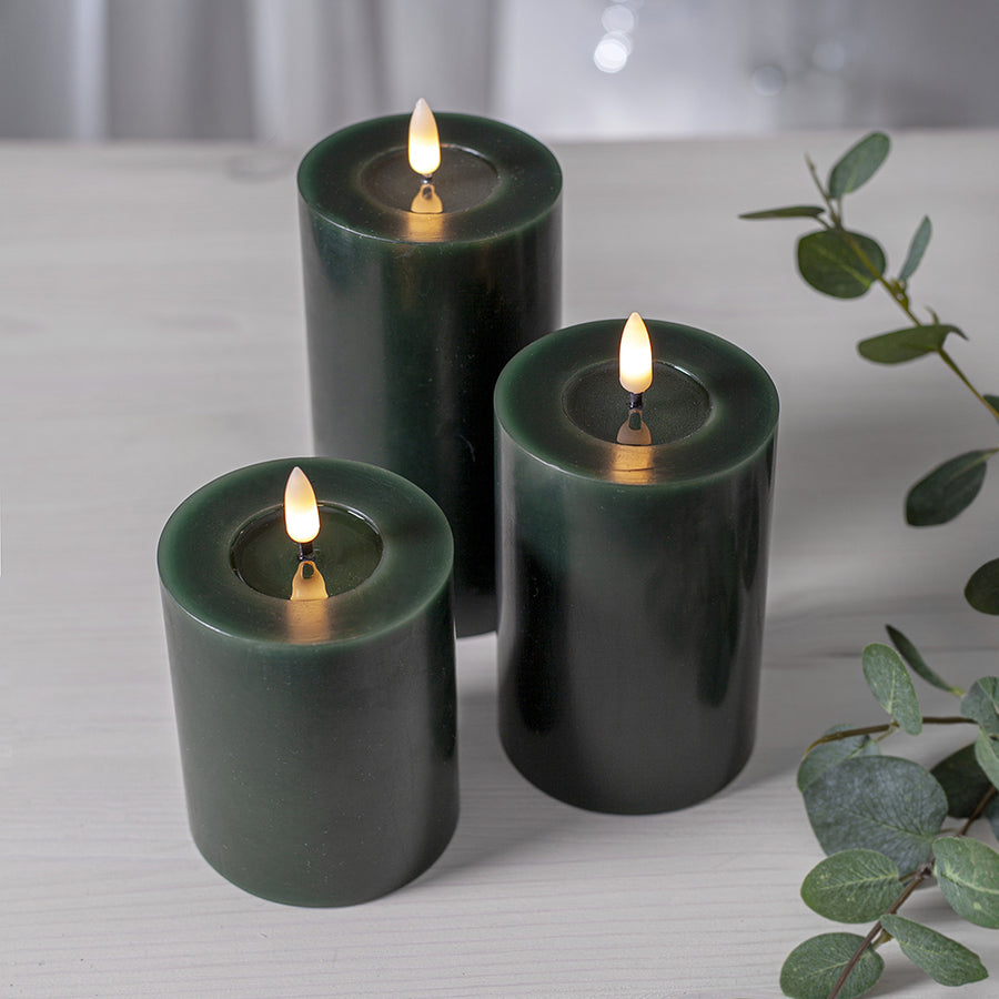 Három különböző méretű zöld LED oszlopgyertya valódi viaszból, meleg fényű pislákoló lánggal, világos fa felületen elrendezve.