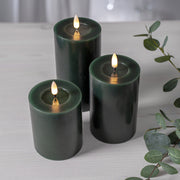 Három különböző méretű zöld LED oszlopgyertya valódi viaszból, meleg fényű pislákoló lánggal, világos fa felületen elrendezve.