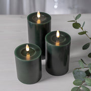 Három különböző méretű zöld LED oszlopgyertya valódi viaszból, meleg fényű pislákoló lánggal, világos fa felületen elrendezve.