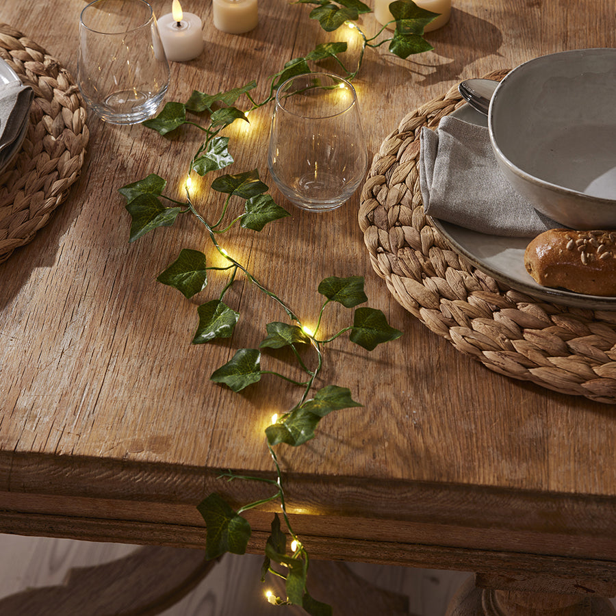 Megterített, rusztikus faasztalon elhelyezett, világító Hedera LED-es mű borostyán girland, meleg fehér fényű LED-ekkel és gyertyafényes hangulattal kiegészítve.