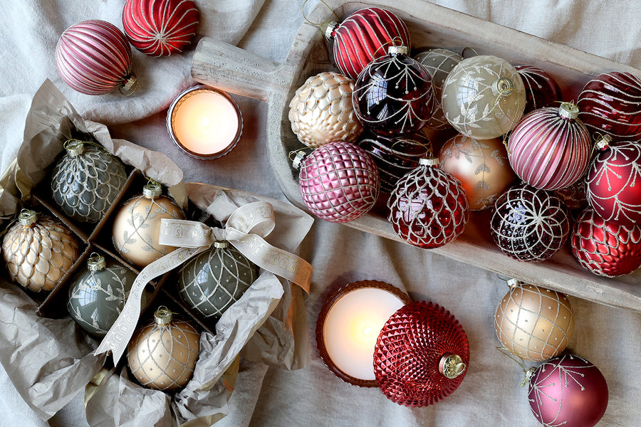 Heritage karácsonyi üvegdíszek pezsgő és bordó árnyalatban, gyertyafényes enteriőrben elhelyezve.