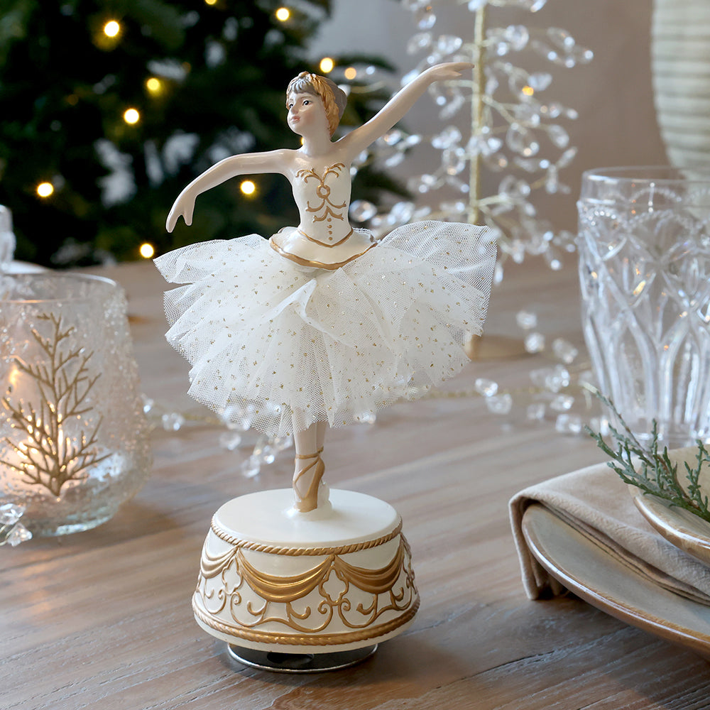Chic Antique balerina figura karácsonyi terítékben, üveg mécsestartó és poharak mellett, fényfüzéres karácsonyfa előtt.