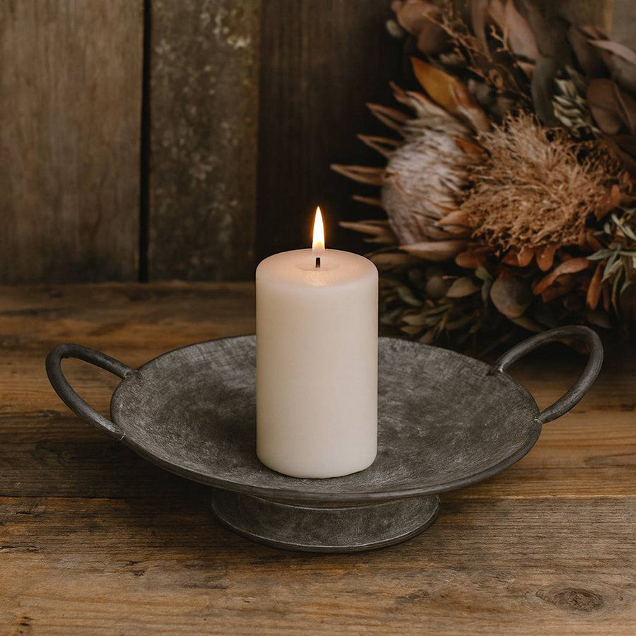 Égő gyertya a JUNKER szürke bádog tálban rusztikus fa felületen, szárított virágkompozícióval a háttérben.