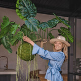 Nő által tartott nagyméretű Monstera Deluxe függeszthető mű könnyezőpálma dekoráció, trópusi hangulatot teremtve