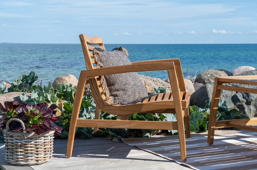 Közelkép egy teakfa karosszékről szürke díszpárnával, háttérben tengeri növényzettel és sziklás parttal.