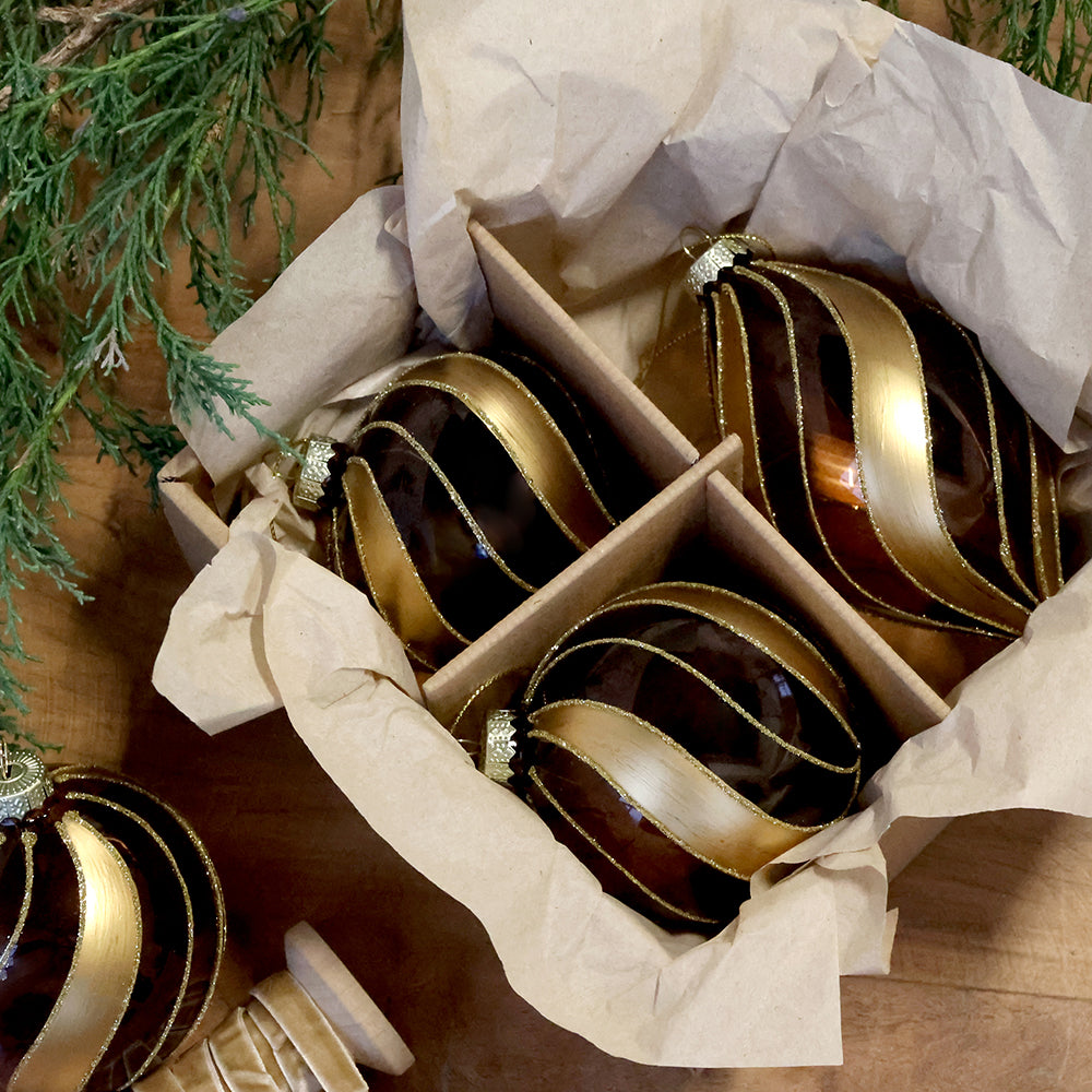 Chic Antique mokka-arany üvegdíszek papírral bélelt dobozban, fa felületen, örökzöld ágak és aranyszínű szalag mellett.