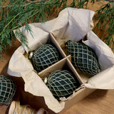Chic Antique olívazöld hálómintás üvegdíszek papírral bélelt dobozban, fa felületen, örökzöld ágak mellett.