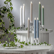 Pislákoló fényű LED vacsoragyertyák poharakban, hangulatos, természetes környezetben, zöld ágakkal körülvéve.