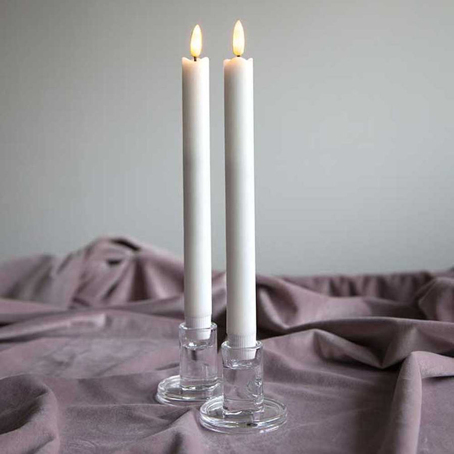 2 darab fehér színű, viaszból készült, meleg fehér fényű led gyertya üveg gyertyatan, bézs színű asztalterítőn, szürke fal előtt 