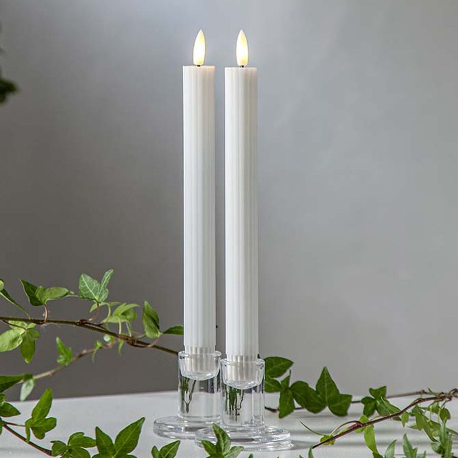 Fehér LED vacsoragyertyák dekor pohárban, meleg fényű lánggal, természetes zöld díszítéssel és elegáns hangulatban elrendezve.